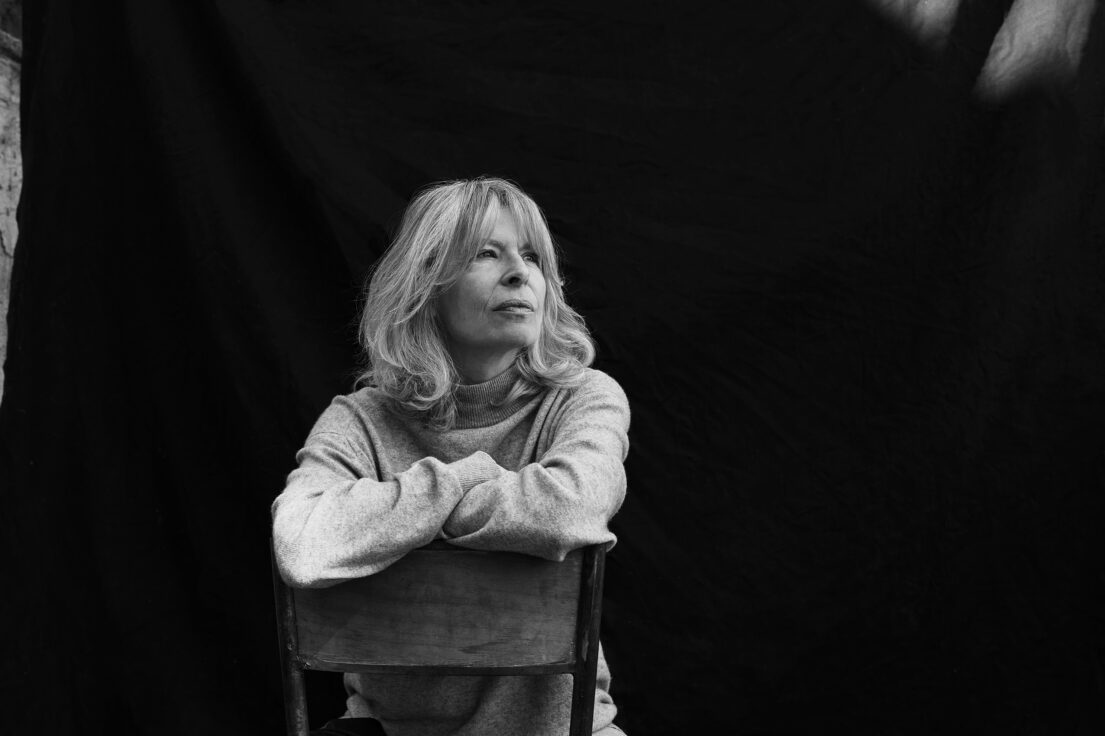 A contemplative woman with crossed arms sits on a chair against a dark backdrop.