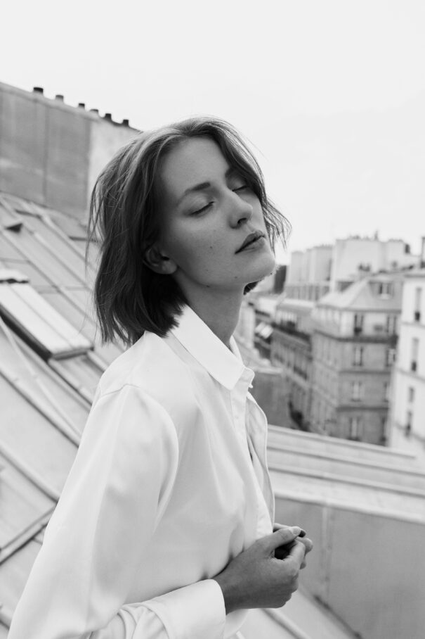 A woman stands on a rooftop with her eyes closed, wearing a white shirt, amidst an urban backdrop.