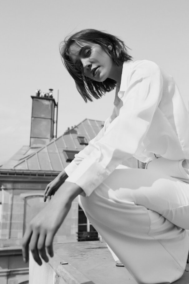 A contemplative woman in white attire crouches on a rooftop with classic French architecture.