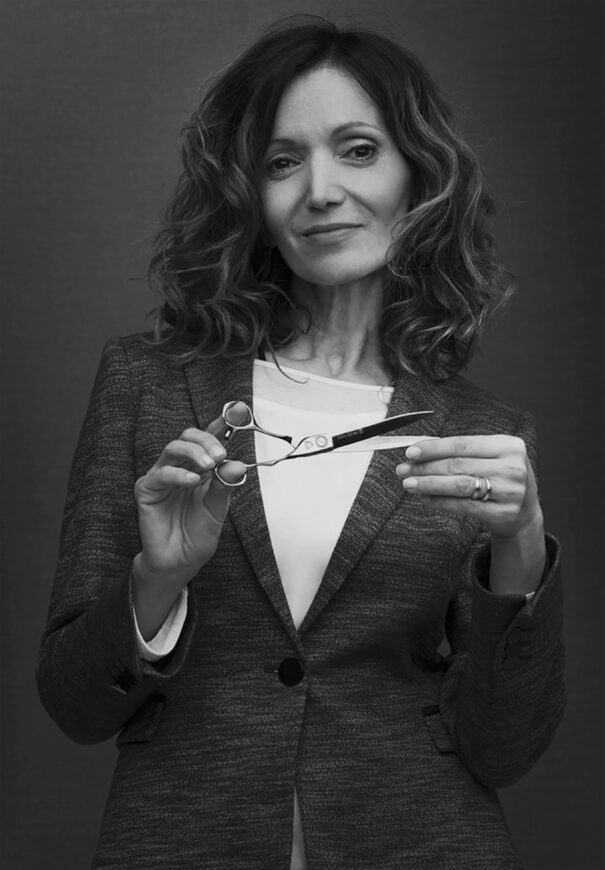 Leatitia Guenaou en costume portant une chemise blanche, avec les cheveux bouclés, tient des ciseaux en position de coupe.