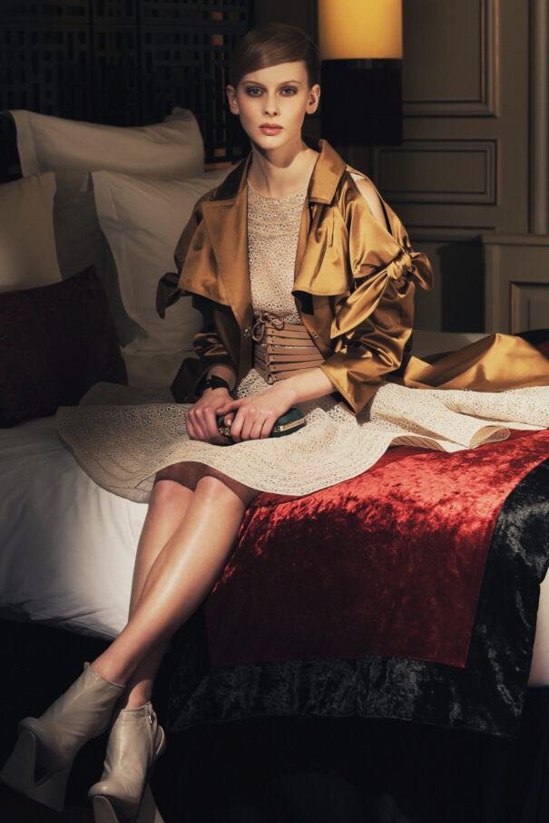 Woman in golden jacket and beige dress sits elegantly on a bed with red blanket.