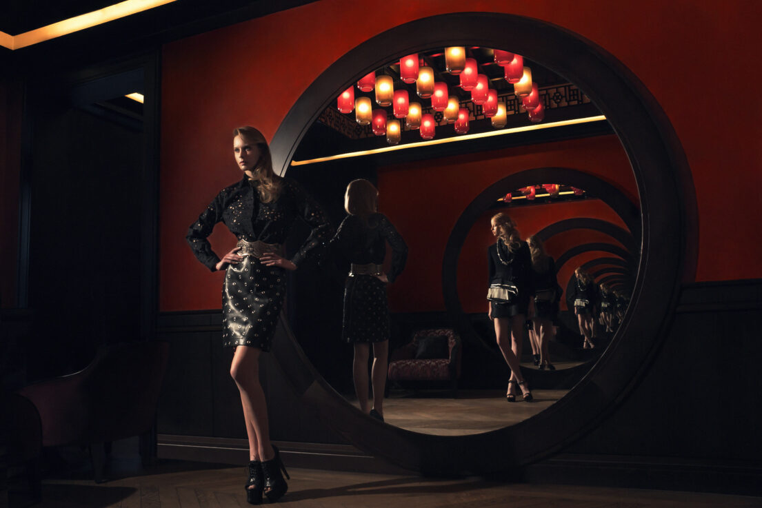 Woman in black dress with high heels stands confidently in front of circular mirrors reflecting her image, set against red walls and warm lighting.