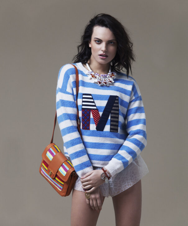 Woman in blue and white striped sweater with colorful "M" design, orange bag, and statement necklace.