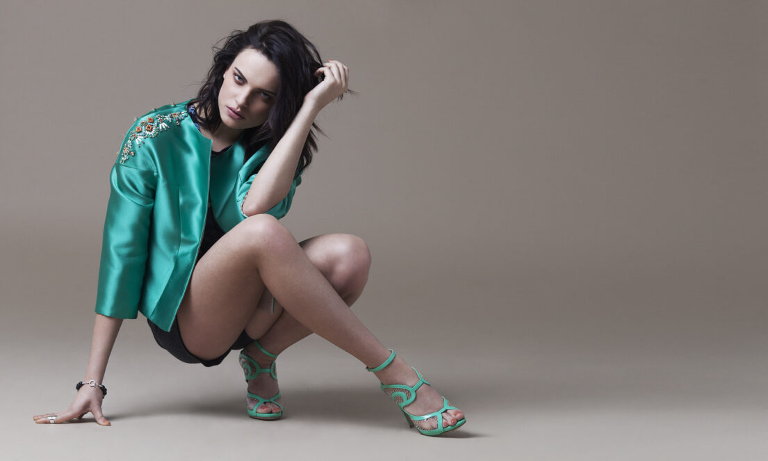 Woman in teal jacket and heels crouching with one hand on the ground and the other touching her hair.