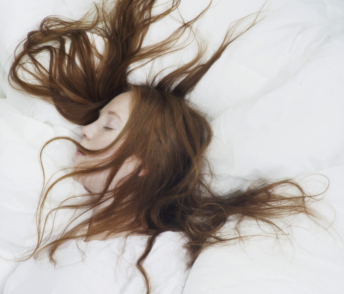 A Woman with long red hair is lying down with eyes closed, surrounded by white bedding.