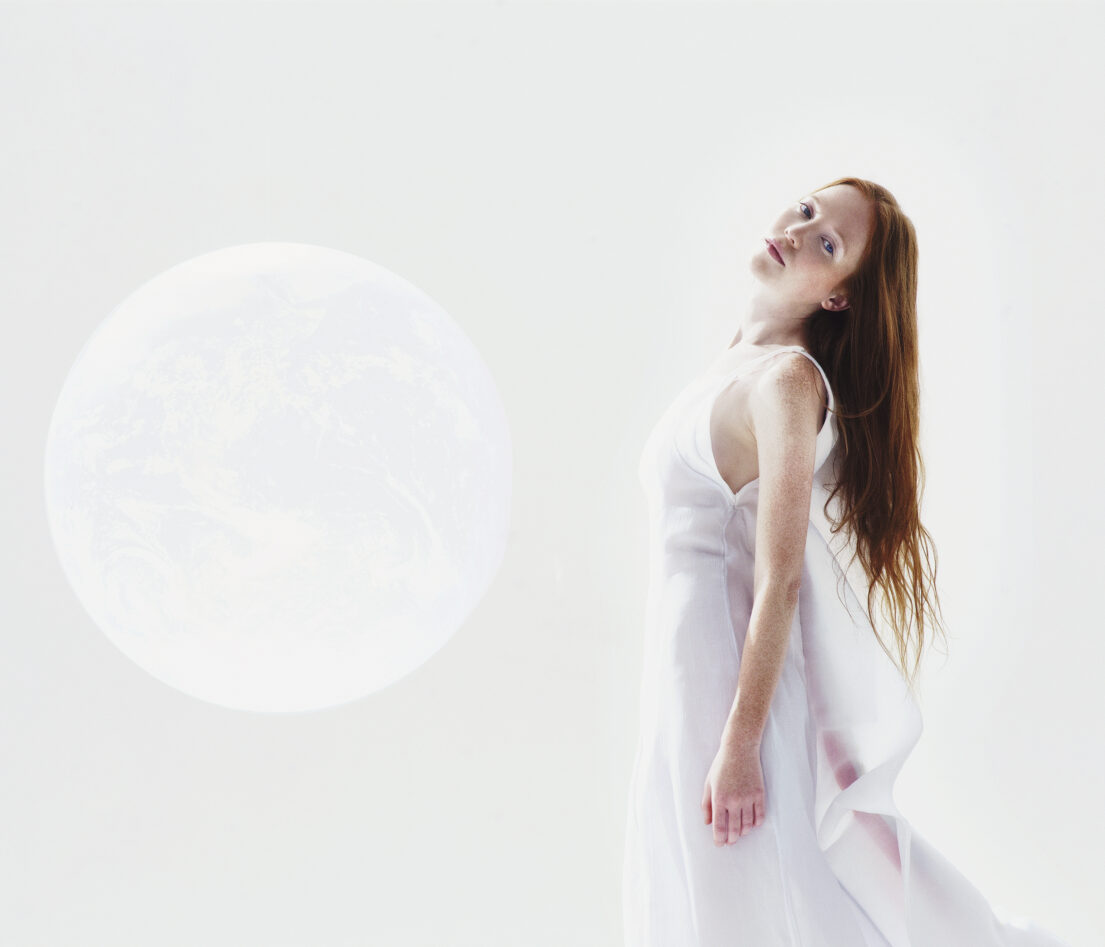 A woman in white dress with flowing hair stands before an ethereal moon backdrop.
