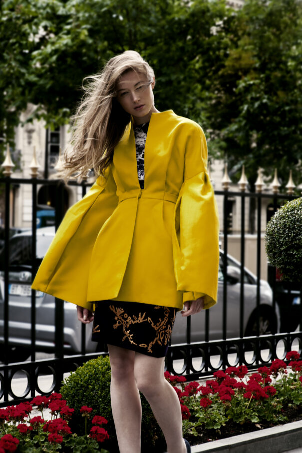 Woman in yellow coat with black floral skirt, walking past red flowers and greenery.