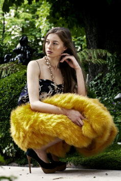 Woman in black floral dress with yellow fur wrap, sitting outdoors among greenery.