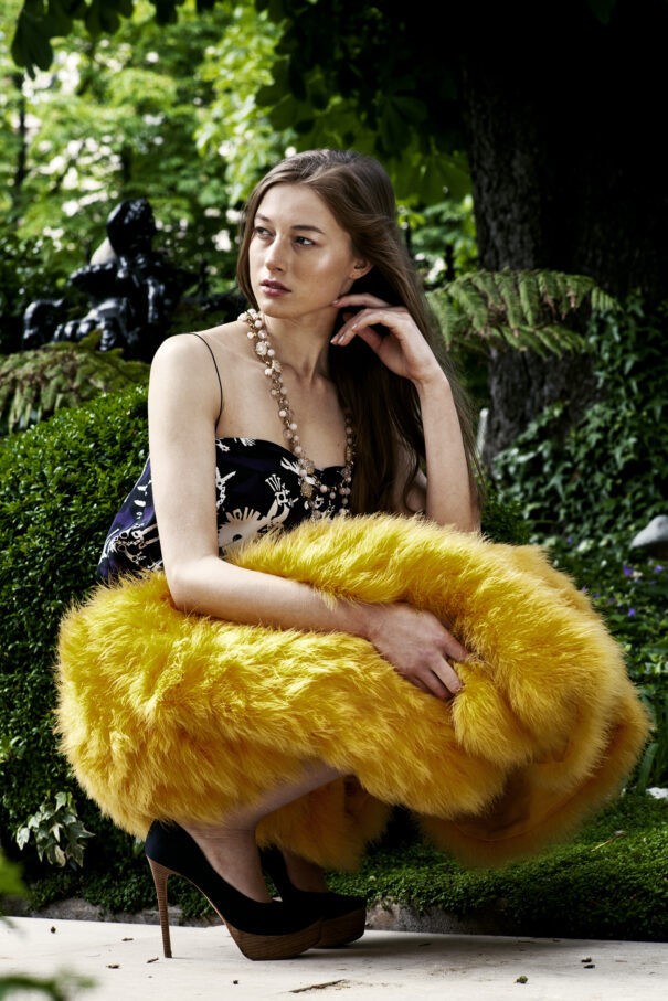Woman in black floral dress with yellow fur wrap, sitting outdoors among greenery.