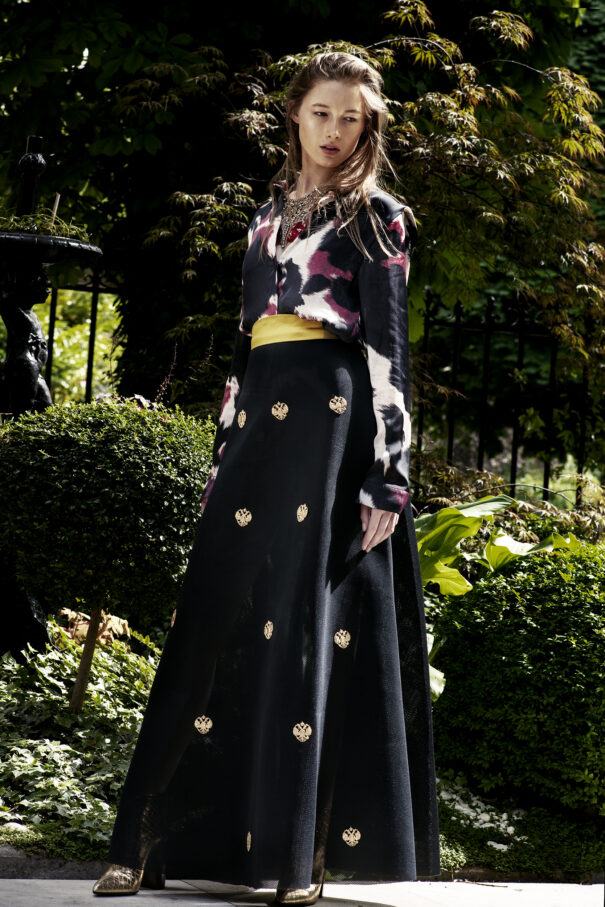 Woman in floral-patterned top and black skirt with gold accents, standing amidst greenery.