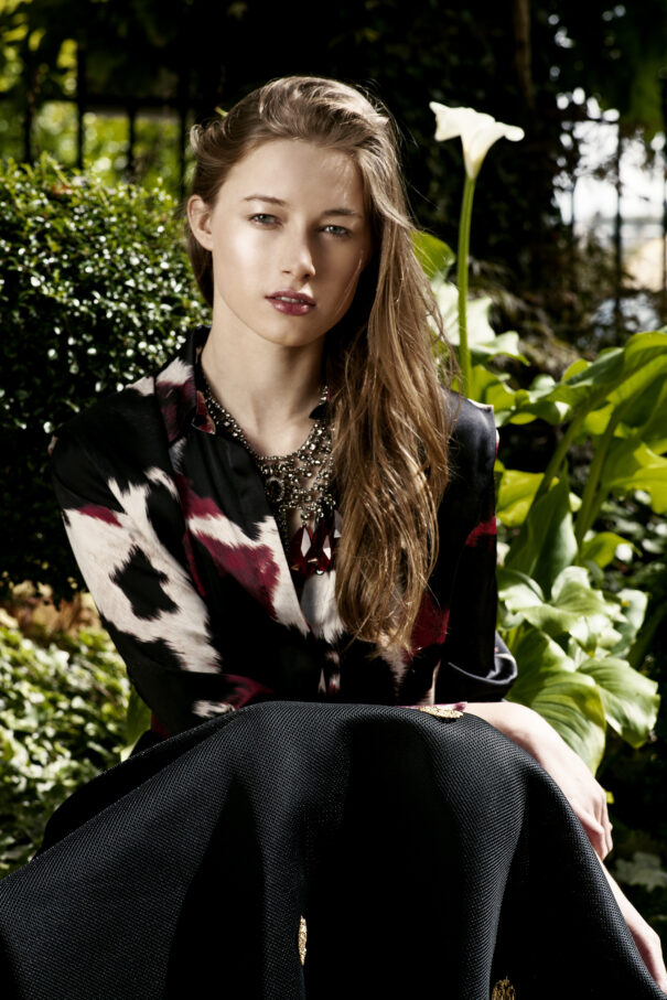 Woman with long hair wearing a patterned blouse and black skirt, sitting outdoors among greenery.