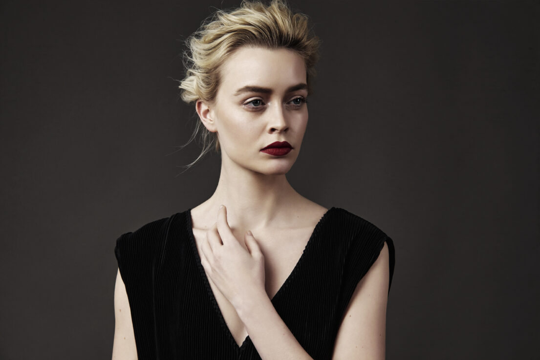 Blonde woman with red lipstick wearing black dress, hand on chest, against dark background.