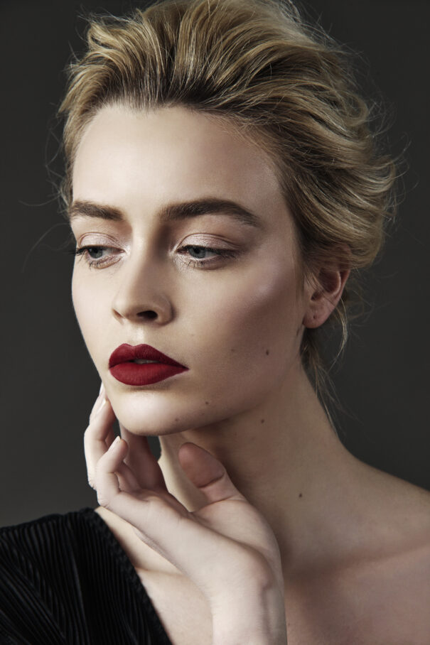 Blonde woman with red lipstick, elegant makeup, and poised hand gesture against dark background.