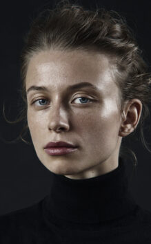 Portrait beauté de Kate Zubok, Une femme avec des cheveux bruns attachés, regard direct, sourcils épilés, regard bleu, maquillage minimaliste, en col roulé noire, fond noir.