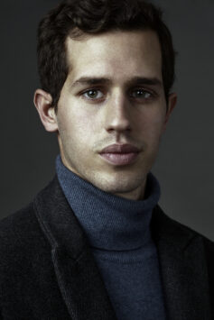 Victor Belemondo, Man with dark hair and eyes, wearing a blue turtleneck sweater and black coat against a gray background.