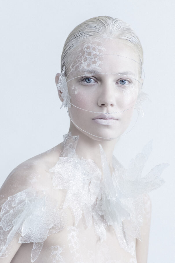 A woman with pale skin and blue eyes adorned with intricate white lace-like patterns on her face and shoulders.
