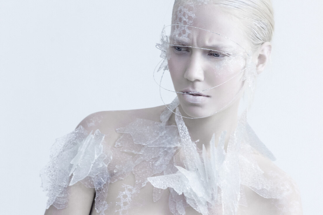 A woman with pale skin and blonde hair is adorned with intricate white lace-like patterns on her face and shoulders, set against a plain background.