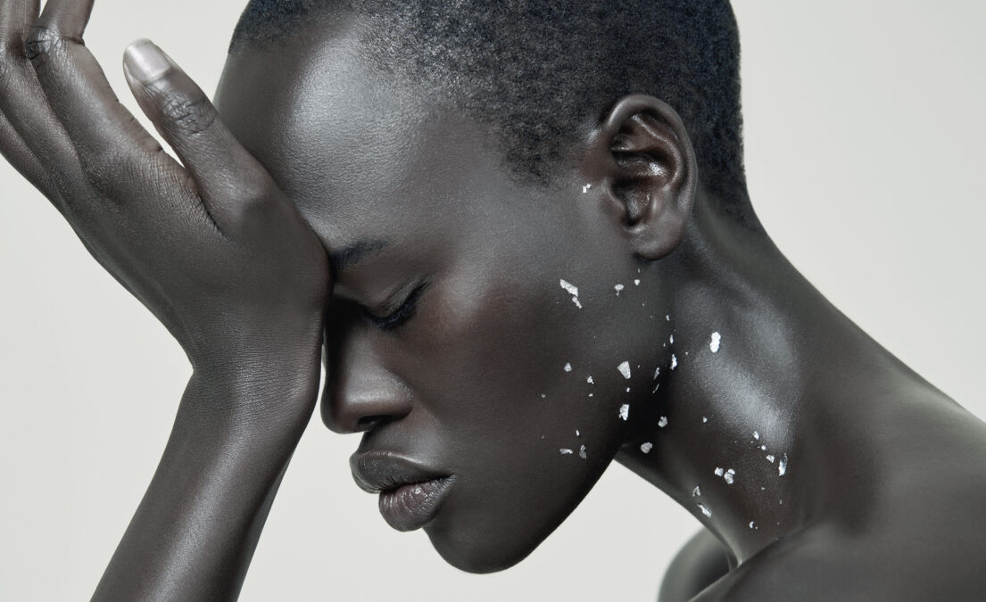 A contemplative individual with short hair, closed eyes, and white speckles on their skin, hand resting on forehead.