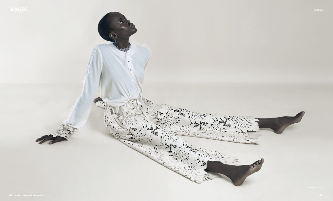 A Woman sits on the floor with legs extended, wearing a white blouse and patterned pants, looking upwards.