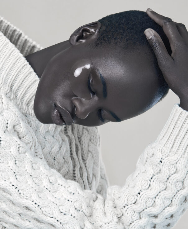 A Woman with short hair wearing a textured white sweater, eyes closed, hand on head.
