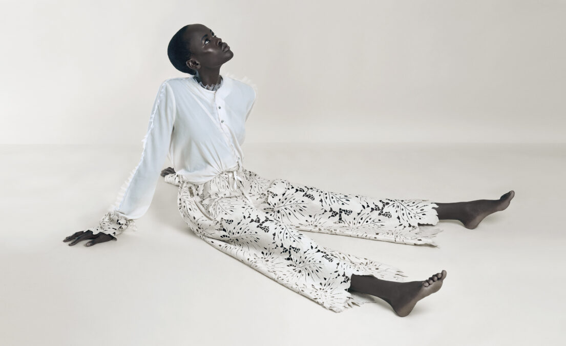 A Woman sits on the floor with legs extended, wearing a white blouse and patterned pants, looking upwards.