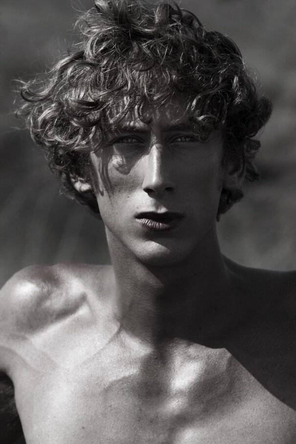 Curly-haired man with intense gaze, shirtless, dramatic lighting.