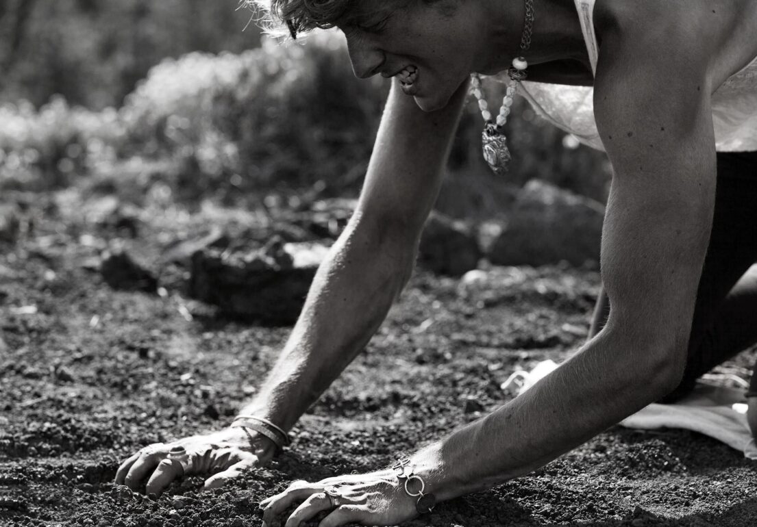 A Man kneels on the ground with his hands touching the soil, wearing bracelets and a necklace.