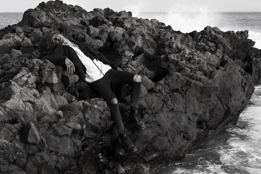 Man reclining on jagged rocks by turbulent sea.