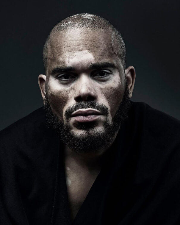 Man with intense gaze, dark clothing, and visible facial hair against a dark background.