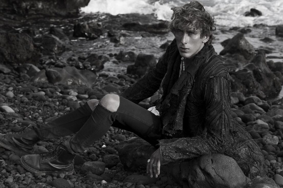 A contemplative individual sits on a rocky shore, dressed in dark attire with waves crashing behind them.