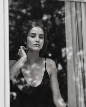 A woman gazes through a window with trees reflected on its surface.