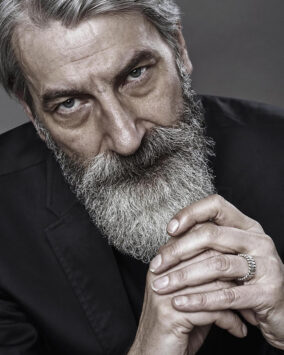 Silver-haired man with beard, wearing a black suit jacket, hands clasped, silver ring visible.