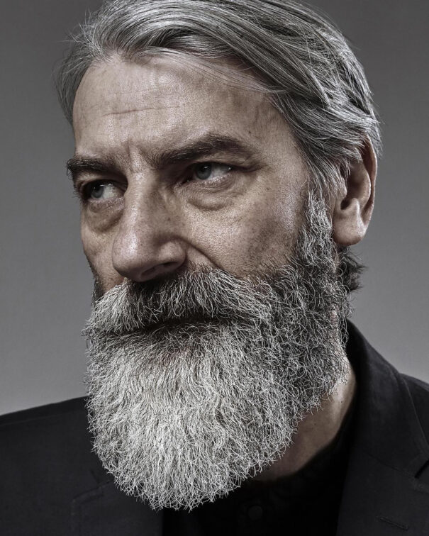 Silver-haired man with full beard, wearing black attire, against gray background.