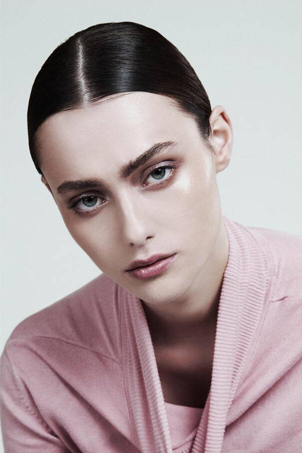 A woman with sleek black hair and green eyes wears a pink cardigan against a white background.