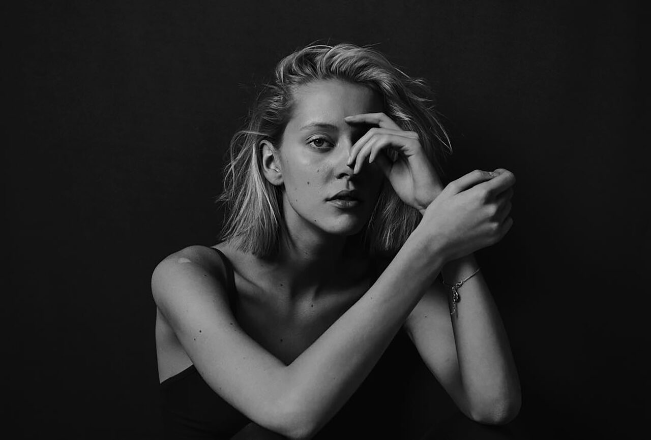 Blonde woman with crossed arms, black tank top, and bracelet, against dark background.