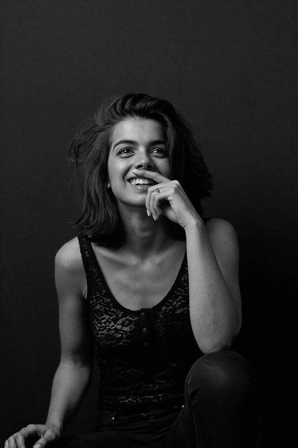 Anais Parello, A woman with short hair smiles while touching her face against a dark background.