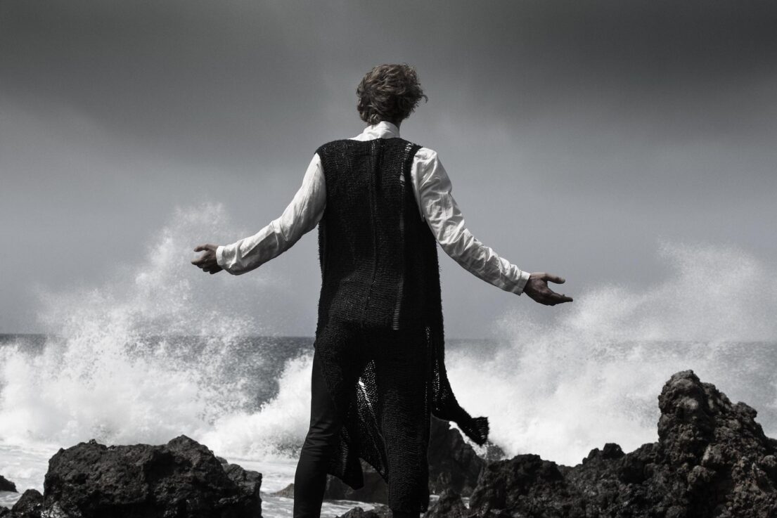 Man with outstretched arms standing on rocks by crashing waves.
