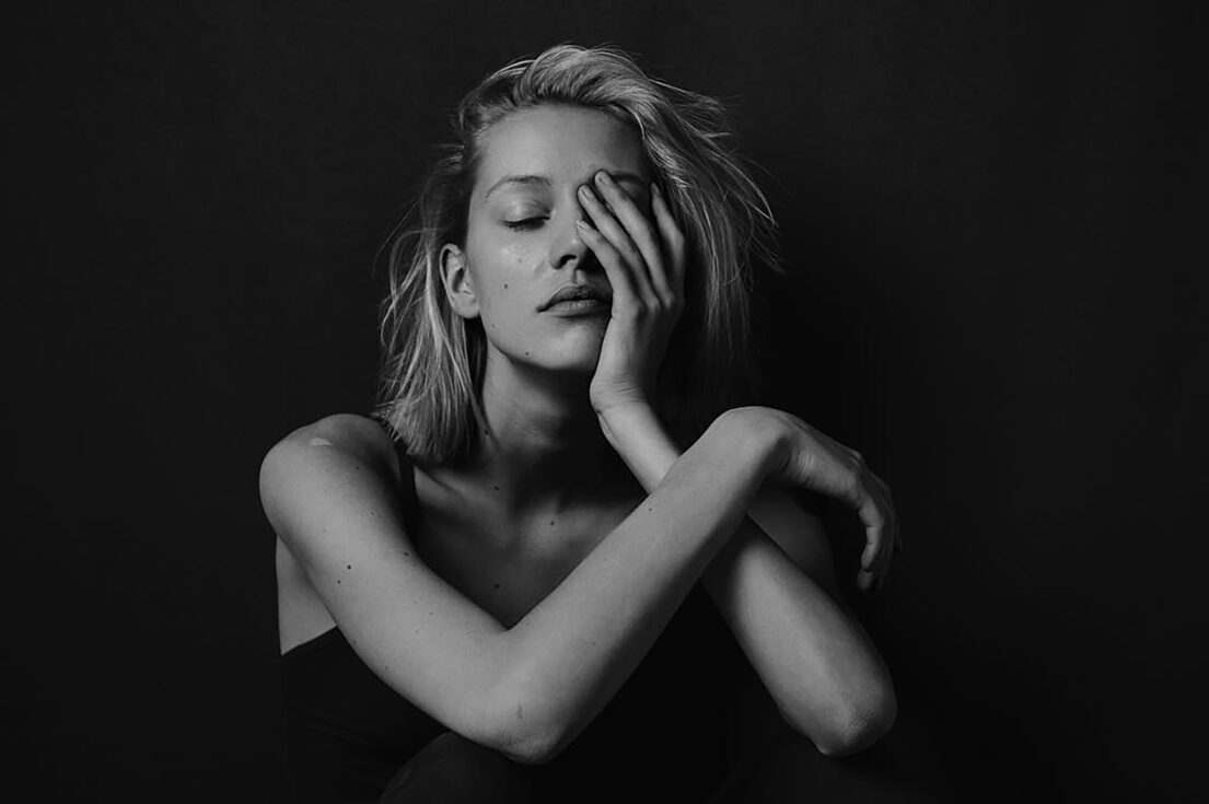 Blonde woman with eyes closed, hand over face, crossed arms, black background.