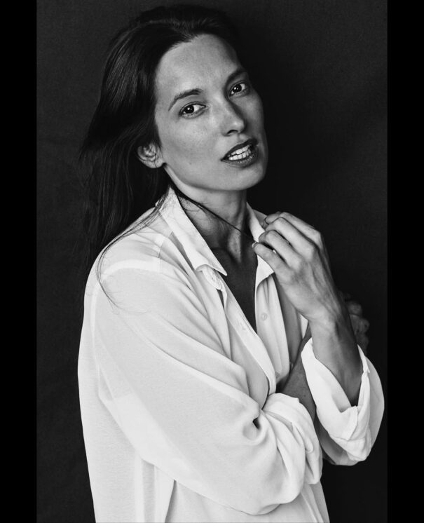 Carole Brana Woman in white shirt with crossed arms, looking thoughtful against dark background.