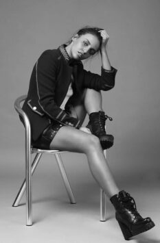Carla Ginola seated on chair, stylish black jacket, shorts, boots, relaxed pose.