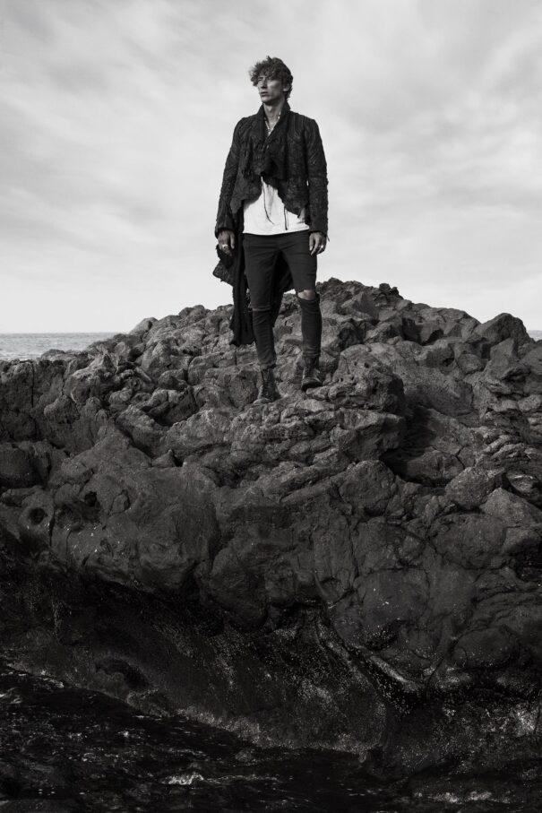 Man stands on rugged rocks by the sea, dressed in dark attire with a dramatic sky backdrop.