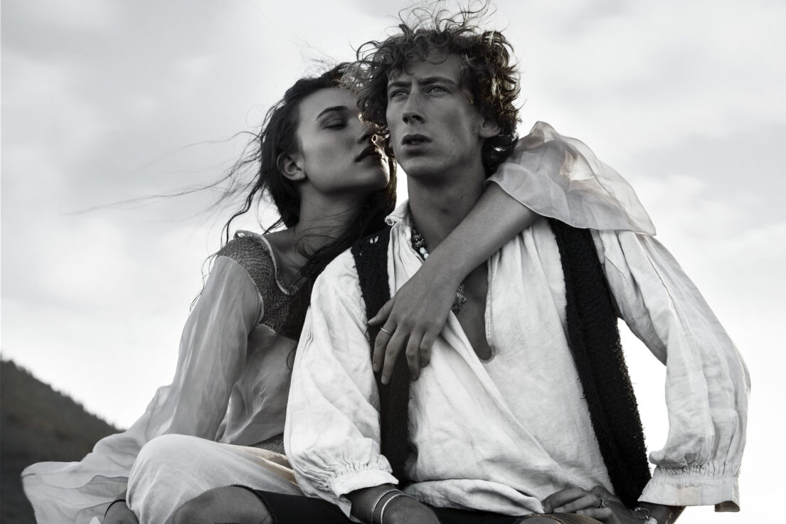 A woman embraces a man from behind, both dressed in period attire against a cloudy sky.