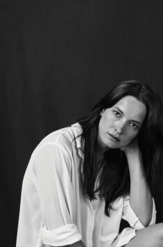 Carole Brana,with long hair wearing a white shirt, resting her chin on her hand against a dark background.