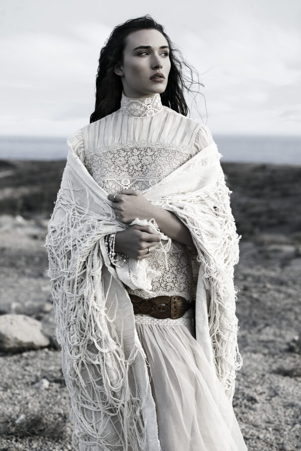 Woman in white lace dress with shawl, standing alone in a desolate landscape.