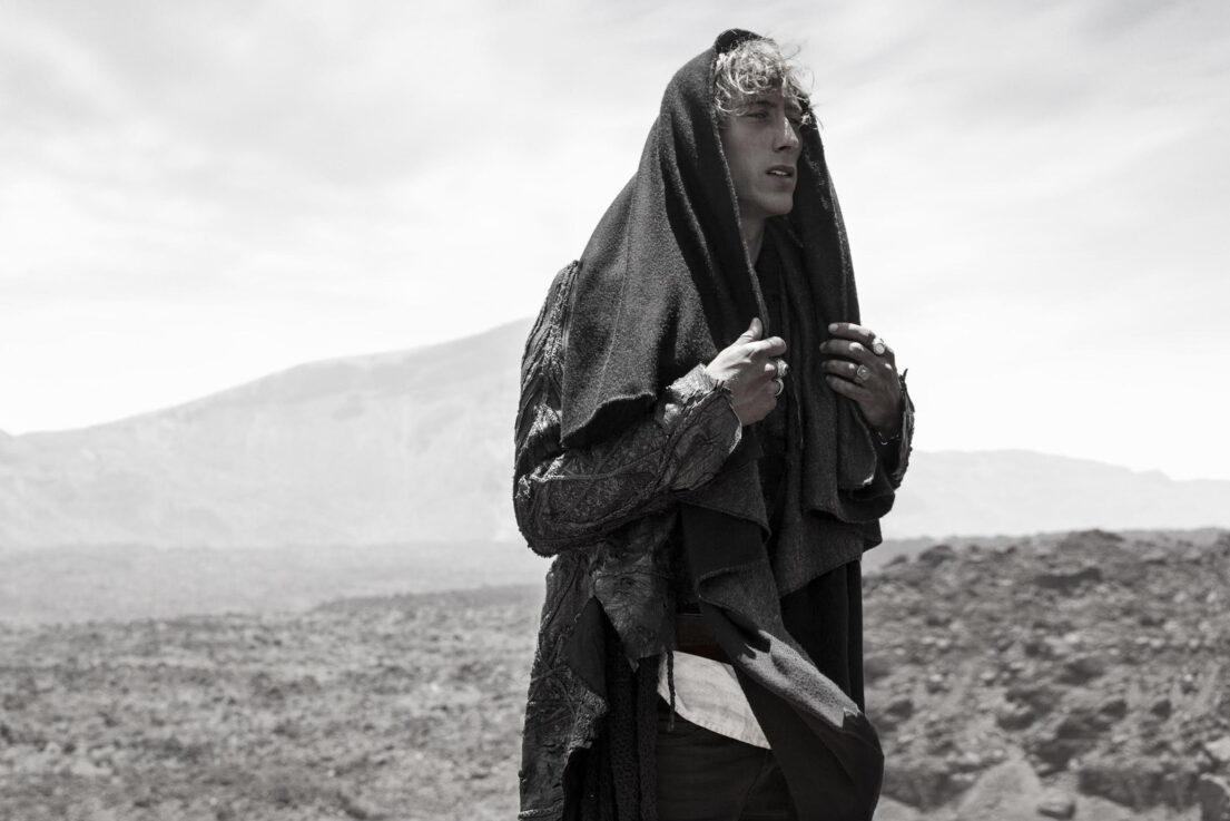 Man in tattered cloak with mountainous backdrop.