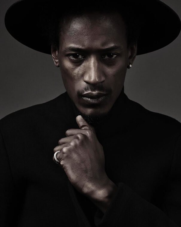 Man with black attire and hat, hand near chin, silver ring, intense gaze.