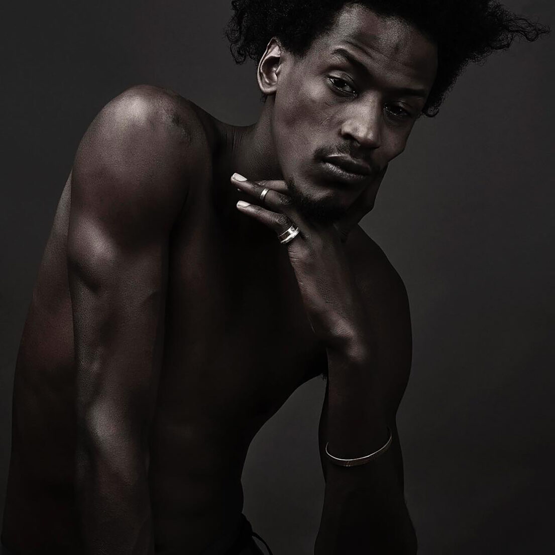 A contemplative man with a muscular build, wearing rings, against a dark background.