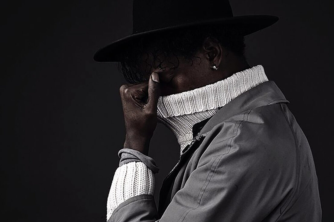 Man in hat and coat with raised collar, hand near face.