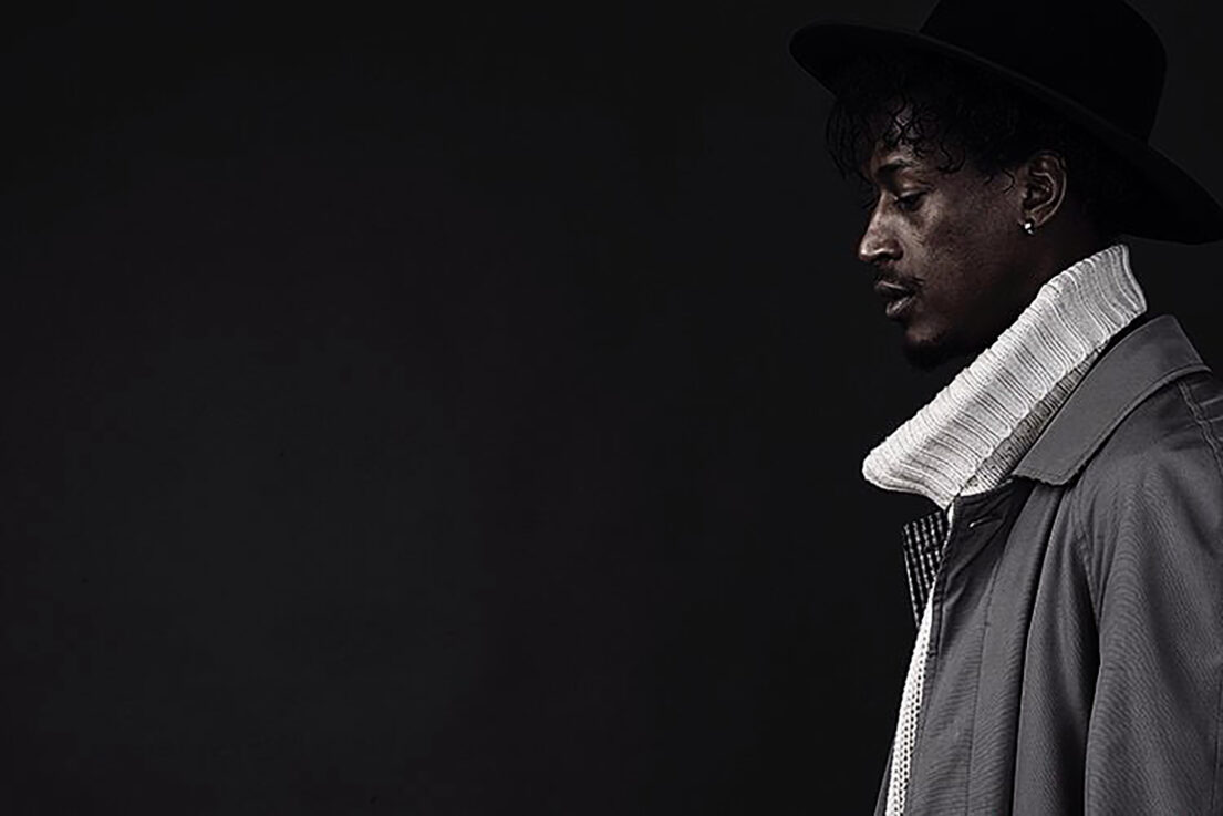 Man in black hat and coat with white collar, side profile against dark background.