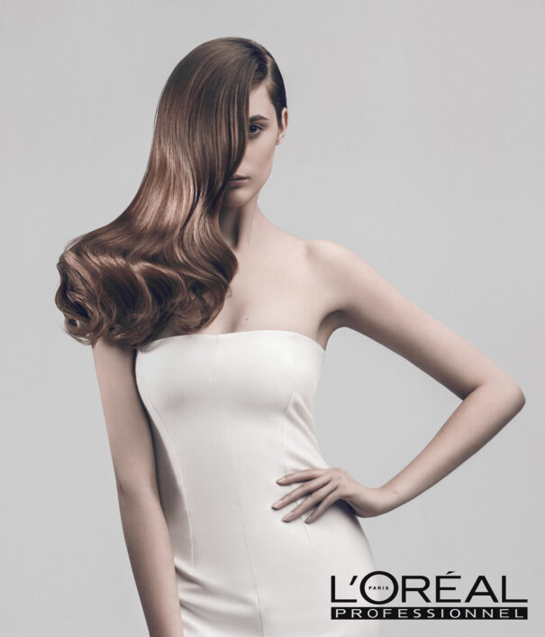 Une femme avec de longs cheveux bruns soyeux, vêtue d'une robe blanche, pose pour une campagne publicitaire L'Oréal Professionnel.